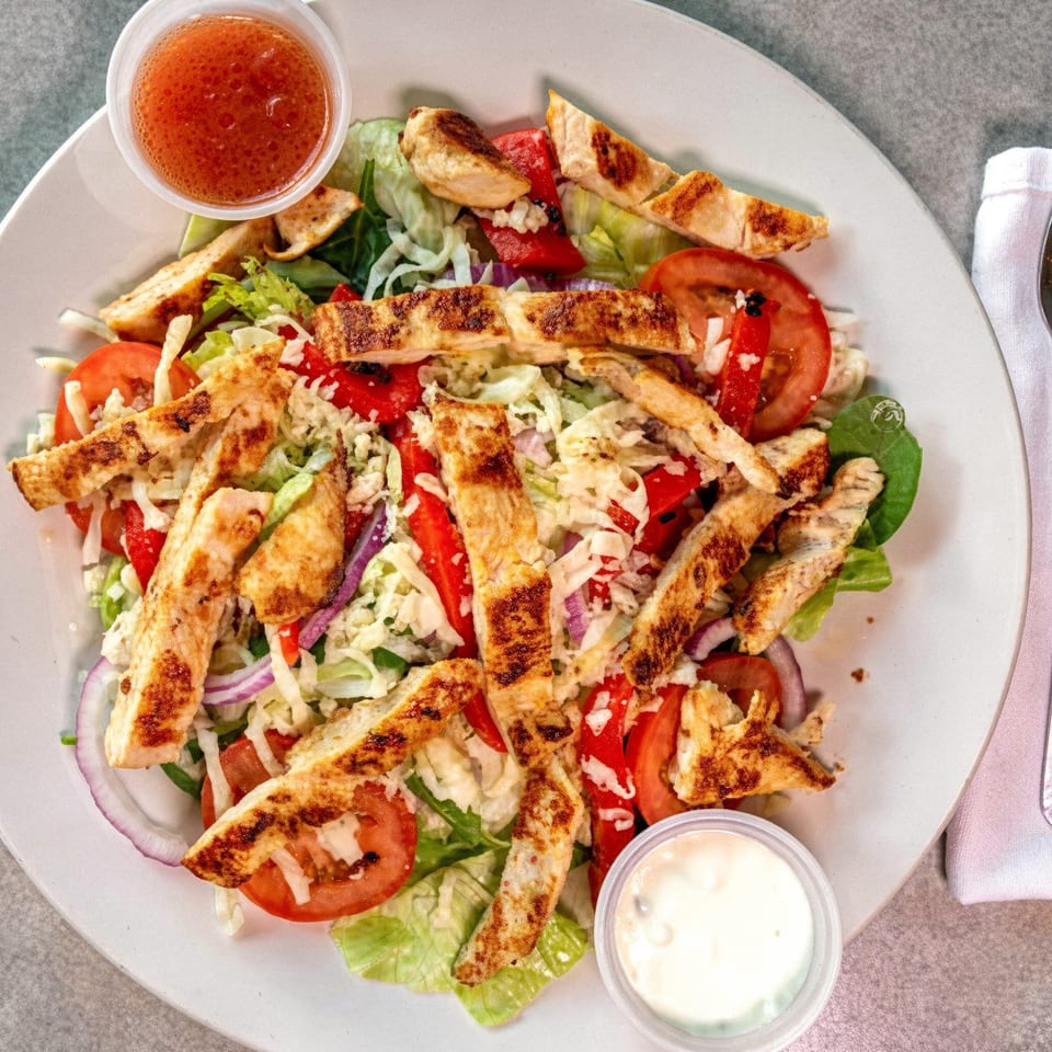 Grilled chicken salad with lettuce, tomatoes, red onions, and two dressings on a white plate.