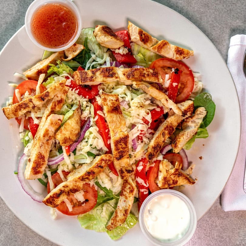 Grilled chicken salad with lettuce, tomatoes, red onions, and two dressings on a white plate.