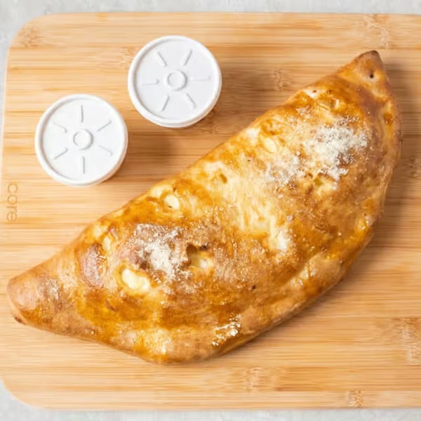 Golden-brown calzone with two white sauce containers on a wooden cutting board.
