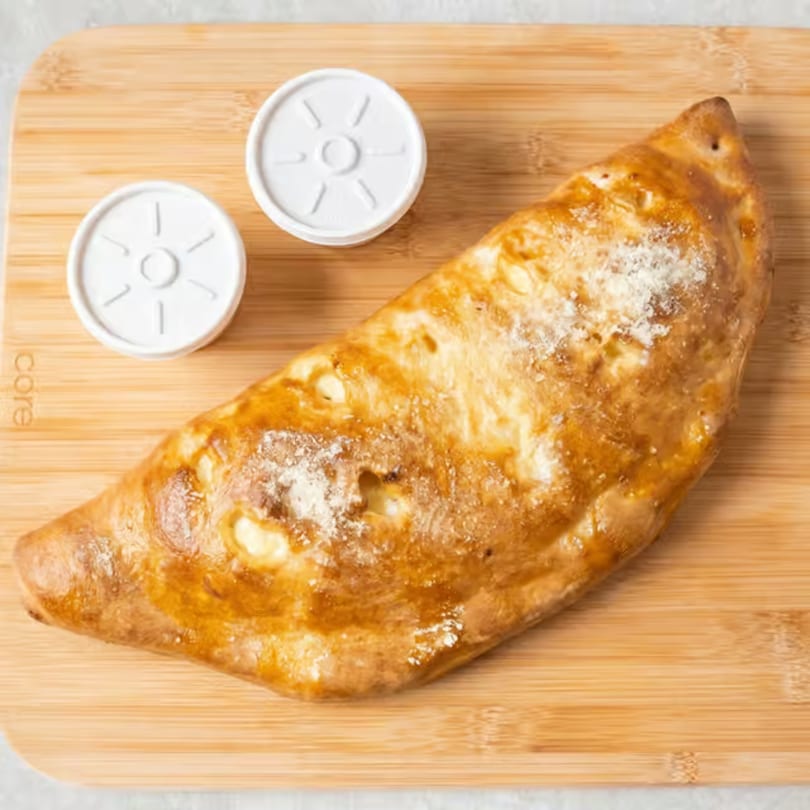 Golden-brown calzone with two white sauce containers on a wooden cutting board.